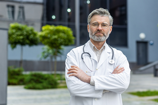 Portrait of senior serious doctor outside modern clinic outdoors, mature man in medical coat looking at camera thinking with crossed arms.