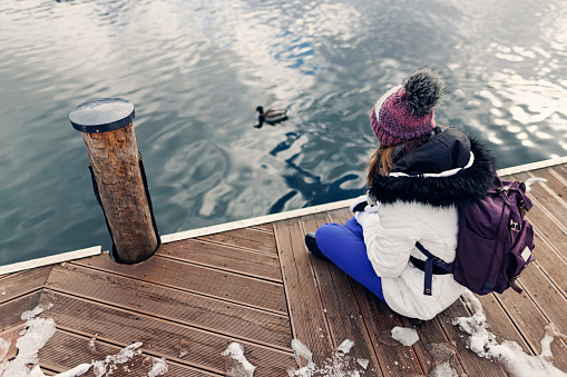 Teenage girl enjoying nature in the European Alps mountains on a winter day. The girl is sitting by the water and looking at a duck swimming by.
Shot with Canon R5