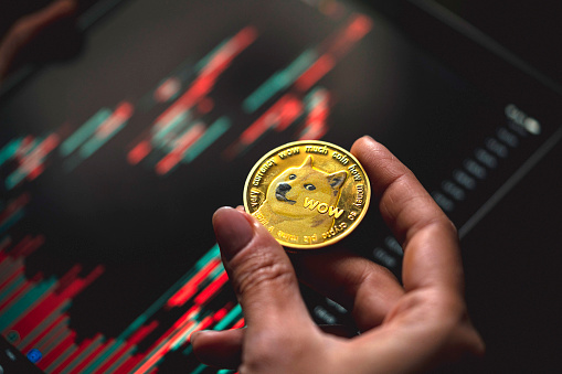 Hand holding a gold Dogecoin cryptocurrency coin with candle stick graph chart and digital background.