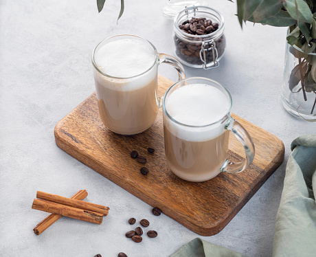 Two cappuccino or latte with milk foam in glasses on a light background with coffee beans, cinnamon and eucalyptus branch. The concept of spring breakfast. Top view.