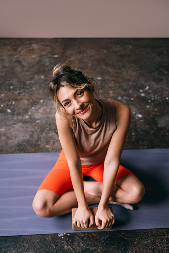 A portrait of a smiling Caucasian athlete in sports clothes sitting on her exercise mat and holding a yoga block while doing her daily workout in the gym.