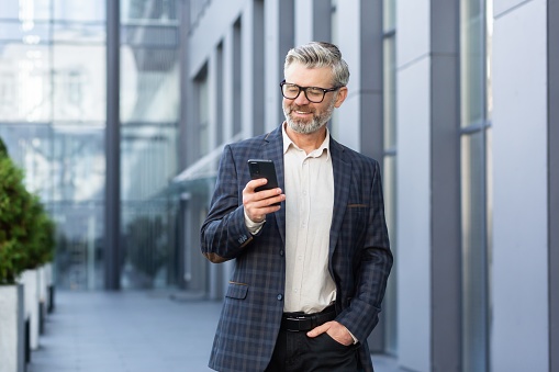 Successful gray-haired businessman walks outside office building, mature boss in glasses uses phone, man smiles and types message, reads online news and browses internet pages