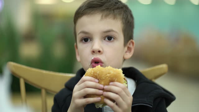 Tired sad boy of seven years old eats a burger at a food court after a school day. Child in sweatshirt enjoying a juicy burger.