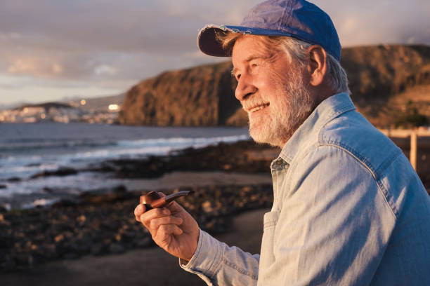 istock Handsome senior bearded man smoking pipe sitting outdoors at the beach at sunset enjoying freedom and sea vacation. Serene retirement concept 1465642427
