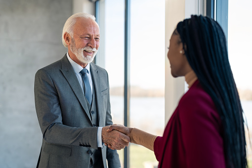 Handsome stylish senior businessman greeting female business partner a successful business woman  with handshake, cheerfully smiling, standing in office near panoramic window. Meeting with coworker.