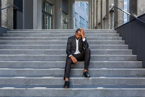 Upset african american man sitting on stairs outside office building, businessman bankrupt in despair, mature boss depressed in business suit outdoors.