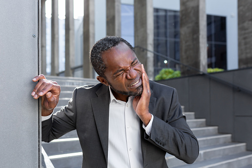 Toothache outside. An African-American man in a suit is standing near an office center, holding his cheek, leaning against a wall. He feels a strong toothache, discomfort in his mouth.