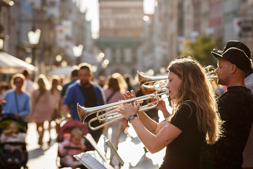 Gdansk, Poland - July 5, 2022: Street musicians playing on brass instruments. Young woman artist plays trumpet in city for crowd of people