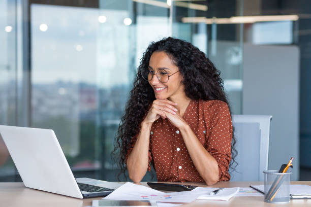istock Young happy and successful businesswoman in glasses working with documents inside office, Hispanic woman with laptop looking at bills and contracts, financier with curly hair using laptop 1465651251