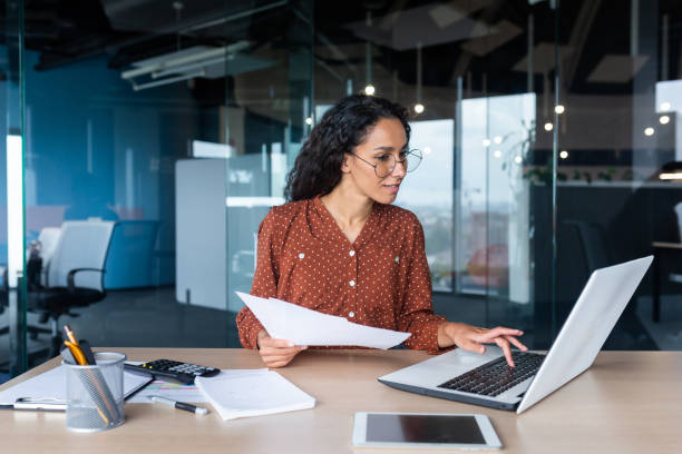 jeune femme d’affaires heureuse et prospère en lunettes travaillant avec des documents à l’intérieur du bureau, femme hispanique avec un ordinateur portable regardant les factures et les contrats, financière aux cheveux bouclés utilisant un ordina - équipement à base de papier photos et images de collection