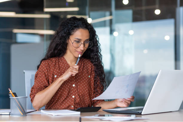 Beautiful hispanic woman in office doing paperwork, successful businesswoman financier working inside office sitting at table using laptop, at work with contracts and bills, thinking about decision