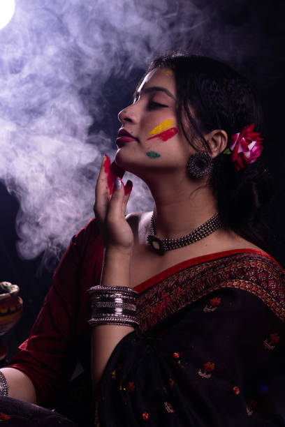 Young Indian girl woman in ethnic attire saree playing with organic dry color powder gulal abeer on face celebrating Holi festival of colors with smoke in the background stock photo
