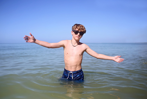 A young teen adolescent boy is looking at the camera with cool attitude while wearing sunglasses and swimming in the ocean on a summer day.