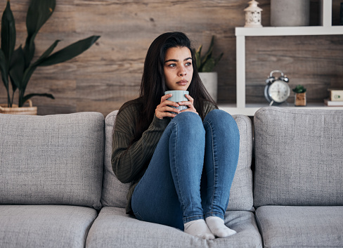 Depression, sad and woman with coffee on a sofa for thinking, lonely and isolated in a living room. Stress, anxiety and girl drinking tea on couch, looking depressed, nostalgic and pensive in a home