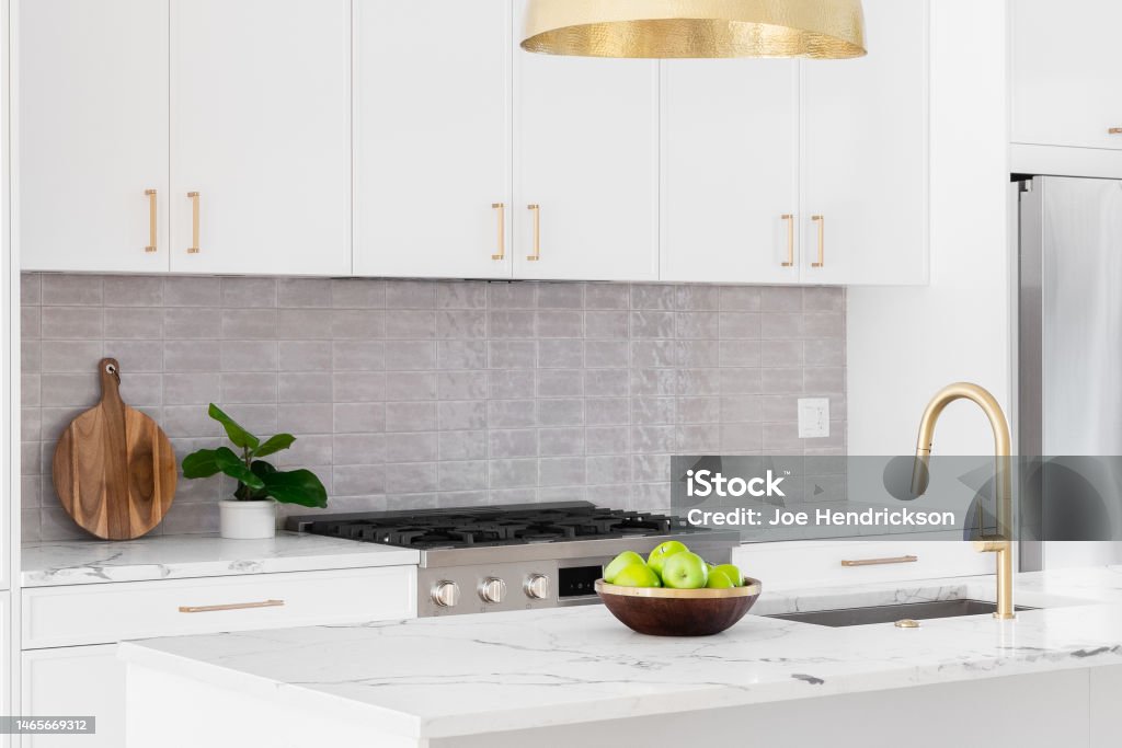 A white kitchen detail with gold light and faucet. A kitchen detail with white cabinets, gold faucet and light hanging over the island, and a tiled backsplash. Kitchen Stock Photo