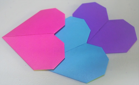 Three traditional origami hearts photographed at an angle with a white background. Hearts are the colors of the bisexual pride flag.