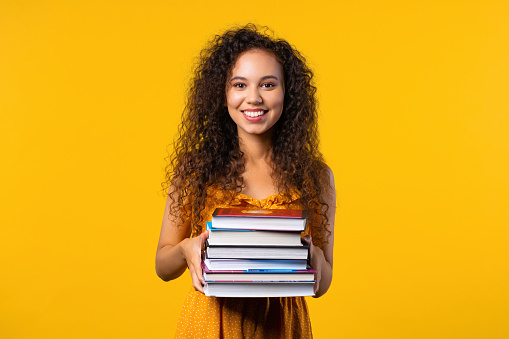 Colorful student woman with stack of books from library on yellow background. Happy girl smiles, she is happy to graduate. High quality photo