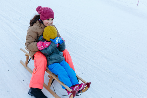 two children sledding on snow slope at one wooden sleigh