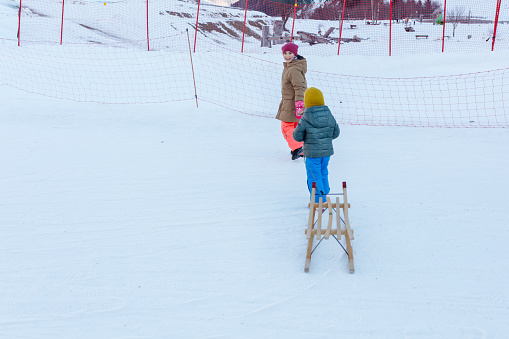 two children pulling the wooden sledge on snow slope in bright clothes