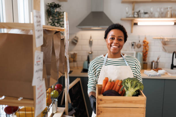Portrait of young beautiful woman with apron holding wooden crate while posing for a shot in kitchen stock photo