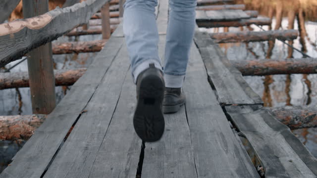 Legs feet of man stepping along suspension bridge over river. Traveler in jeans and leather boots trekking