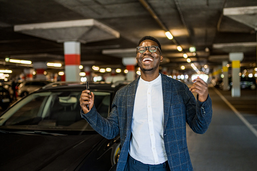 Excited young African American businessman celebrating and laughing while walking in public car garage next to his car.