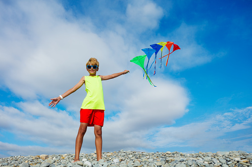 Young smiling boy portrait stand on pebble beach holding many colorful kites in hand over blue sky wearing sunglasses waving hands