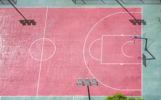 An aerial view of a red basketball court with some silhouettes of the LED vertical spotlights and the basket; view from high above of red and green basketball playground field on a sunny day
