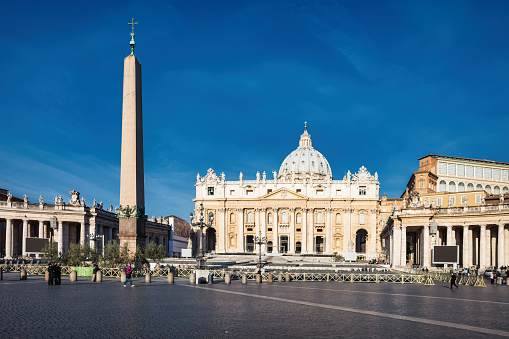 Pedestrians walk in front of St Peter's Basilica on Saint Peter's Square in Vatican on a sunny day.