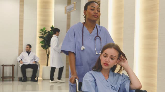 A black nurse pushes a white patient in a wheelchair in a hospital corridor.