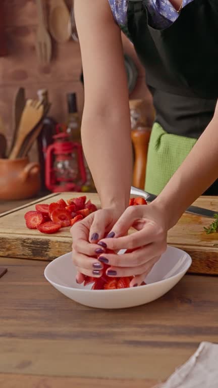 Close-up of woman hands placing slices of strawberries into a bowl to prepare marmalade preserves.
