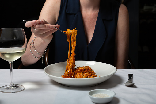 A woman's hands are seen eating angel hair pasta serves with marinara sauce at an up-market restaurant. The pasta is served alongside a glass of white wine and on a white tablecloth.