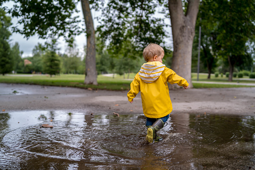 Little boy playing in rainy summer park. waterproof coat and boots jumping in puddle and mud in the rain. Kid walking in summer rain Outdoor fun by any weather. happy childhood