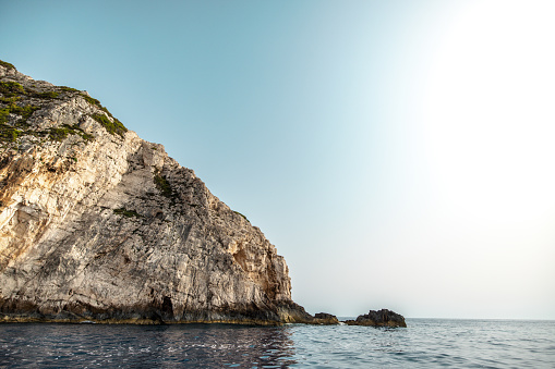 Beautiful high rocky cliffs in Greece on a beautiful sunny day. The sea is calm and the sun is shining. The bottom of the majestic cliff is covered in algae and green moss.