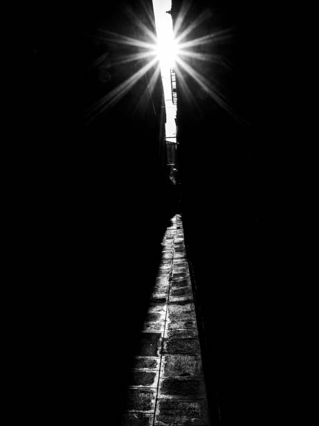 Black and white photograph of the sun shining down a narrow street.