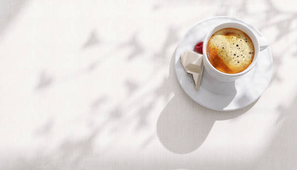 istock Beautiful espresso coffee bubble in white ceramic coffee cup, chocolate, red berry saucer on white tablecloth table with empty space in sunlight, leaf shadow in cafe 1465773029