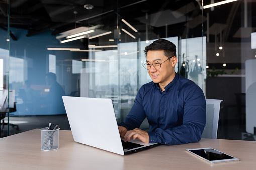 Successful mature asian working inside office using laptop, man typing on keyboard and smiling, businessman in shirt and glasses satisfied with work and achievement results, programmer at work.