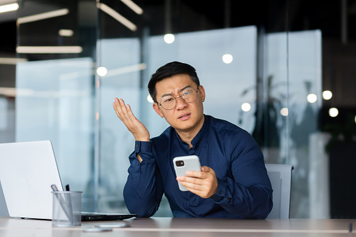 Dissatisfied Asian looking at camera, man inside office holding phone, portrait of dissatisfied businessman at workplace at laptop computer.