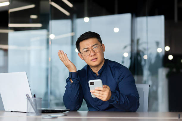 недовольный азиат смотрит в камеру, мужчина в офисе держит телефон, портрет недовольного бизнесмена на рабочем месте за ноутбуком - раздражение стоковые фото и изображения