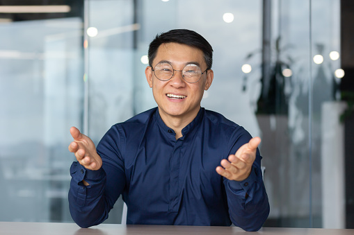 A successful Asian man inside the office is looking at the web camera, talking on a video call with colleagues, gesturing and smiling an online meeting with partners, talking to customers, pov