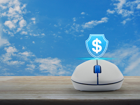 Dollar with shield flat icon on wireless computer mouse on wooden table over blue sky with white clouds, Business money insurance and protection online concept