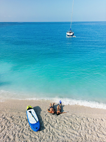 Mature couple enjoying vacation on sailboat and lying and relaxing alone on the beautiful beach. Standup paddleboard is next to them and color of water is amazing. Porto Katsiki beach, Lefkada Island, Greece