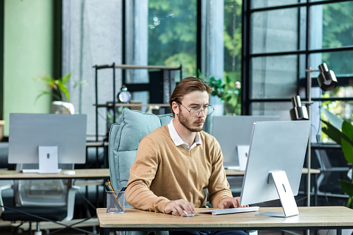 Serious and scorn-centered man in the office at work with a computer, man typing thoughtfully on keyboards, businessman young blond in sweater indoors loft, with green colors, programmer at work.