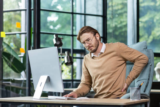 istock Tired businessman at workplace has severe back and lower back pain, young man working sitting at table with laptop and computer in middle of office 1465802889