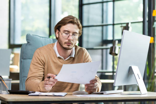 istock A serious young male programmer sits in the office at a table, works at a computer and documents. He holds a project, a plan, bills in his hands 1465803324