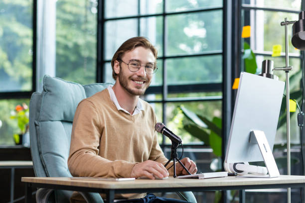 portrait d’un jeune beau professeur. il est assis au bureau du bureau devant le microphone, travaille à l’ordinateur portable, enseigne par appel vidéo. il regarde la caméra, sourit - looking at camera smiling desk isolated photos et images de collection