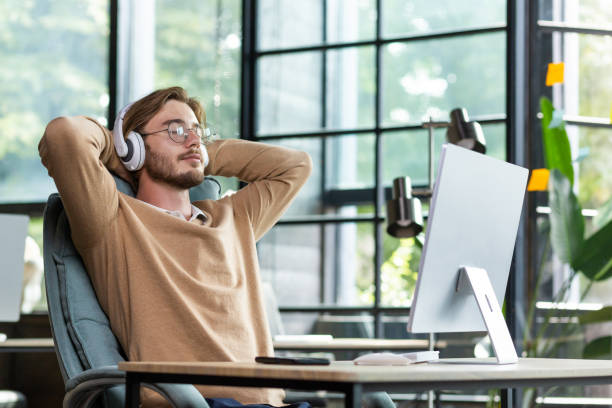 Young successful man in office resting with eyes closed and hands behind head, programmer finished work satisfied with achievement results and listening to music in headphones and podcast online