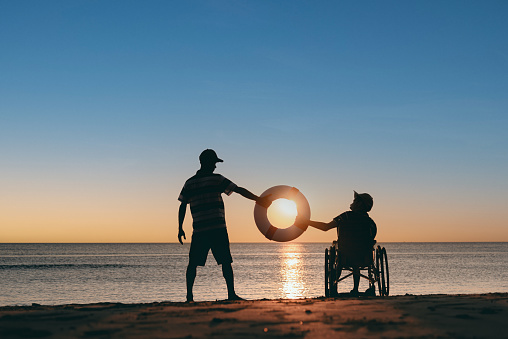 Behind of young man with disability on wheelchair and father or volunteer or caregiver holding rubber ring on the sunset beach, Travel and vacation in summer, Positive photos, mental health concept.