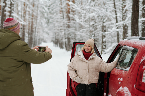 Smiling mature woman in winterwear standing by open door of car and posing for her husband with smartphone taking picture of her