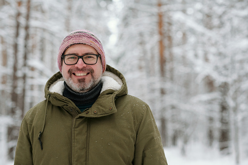 Happy mature man in warm winter jacket with hood, eyeglasses and knitted beanie looking at camera in pinetree forest